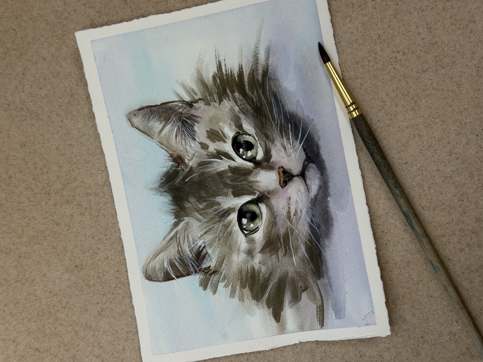 Watercolor kitten portrait with paintbrush beside the paper